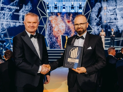Un cadru cu doi bărbați în ținută de gală dând mâna. Bărbatul din dreapta, Maciej Góralski, ține un premiu înrămat cu o emblemă aurie, pe care se poate citi titlul de „Antreprenorul Anului 2022”. În fundalul ușor neclar, se observă o scenă cu structuri geometrice abstracte și motive grafice dinamice în nuanțe de albastru și roșu-portocaliu. În sala de evenimente cu lumină difuză, se pot vedea mai mulți invitați așezați la mese.