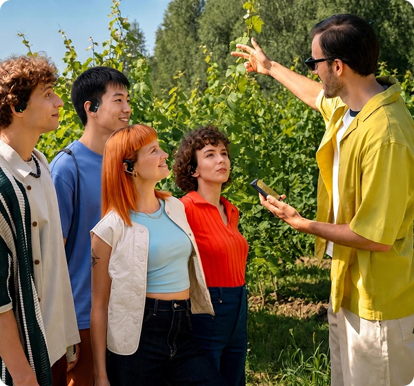Un grup de cinci persoane se află în aer liber, într-o podgorie scăldată de soare. În dreapta, un bărbat îmbrăcat cu o cămașă galbenă și ochelari de soare ține în mână un dispozitiv portabil Vasco Q1 și face un gest spre vița de vie verde. În stânga, patru persoane ascultă cu atenție, fiecare purtând un dispozitiv Vasco E1 negru, demonstrând funcția de conversație în grup în timp real. 