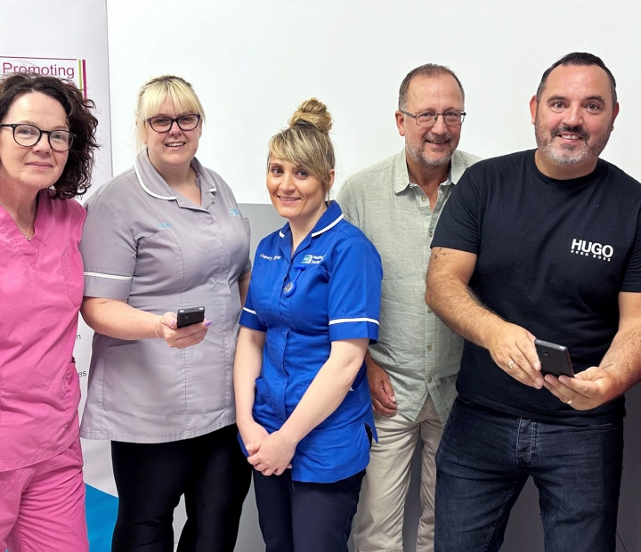 Cinci oameni zâmbitori stau împreună în ținute medicale, doi dintre ei ținând un dispozitiv de traducere.