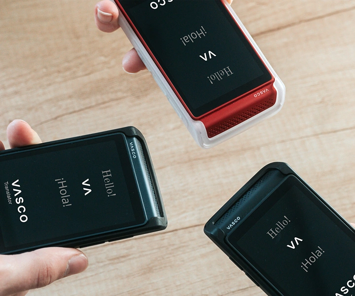 Trei persoane țin în mână dispozitive Vasco Translator Q1 și le apropie între ele; pe ecrane apare ecranul de start cu logo-ul Vasco și mesajele „Hello!” și „¡Hola!”. Dispozitivele sunt prezentate în huse din silicon, în trei culori: Solid Black, Dark Caramel și Milky White.