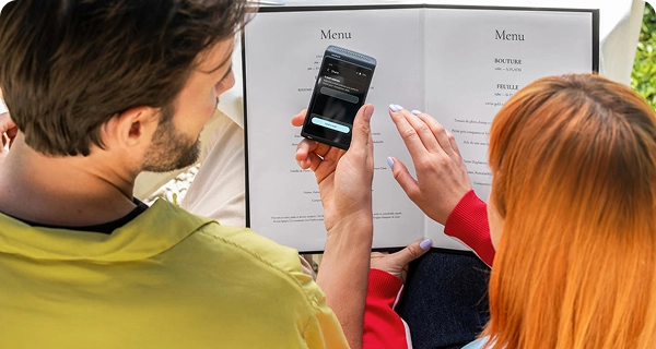 O imagine din spate a unui bărbat și a unei femei cu părul de un portocaliu strălucitor, așezați la o masă de restaurant, uitându-se la un meniu. Bărbatul ține în mână un dispozitiv negru Vasco Translator Q1, pe ecranul căruia apare opțiunea „Share” (Partajare), cu un câmp de introducere a datelor pentru a trimite traducerea prin e-mail. În fundal se văd o față de masă albă și decorul restaurantului.